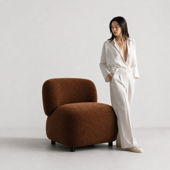 Woman in a white outfit standing next to a brown armchair on a plain background