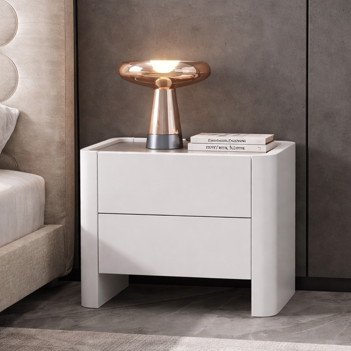 White nightstand with a lamp and books next to a bed in a bedroom setting.