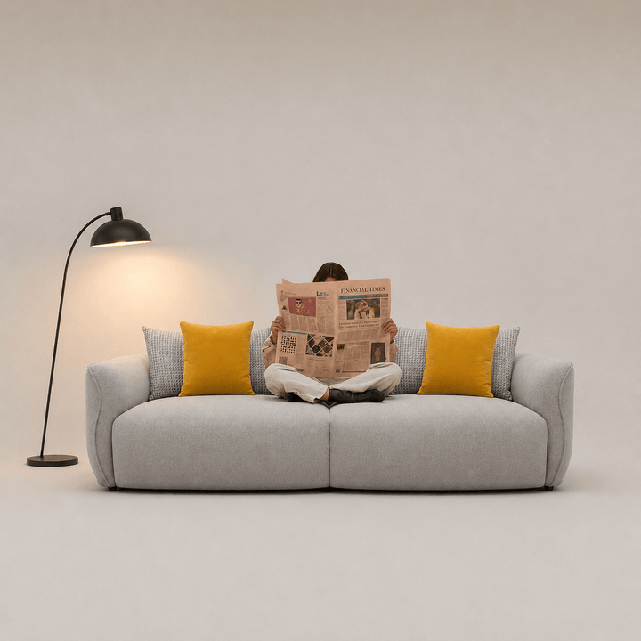 Person reading a newspaper on a gray sofa with yellow cushions, in a minimalistic room.