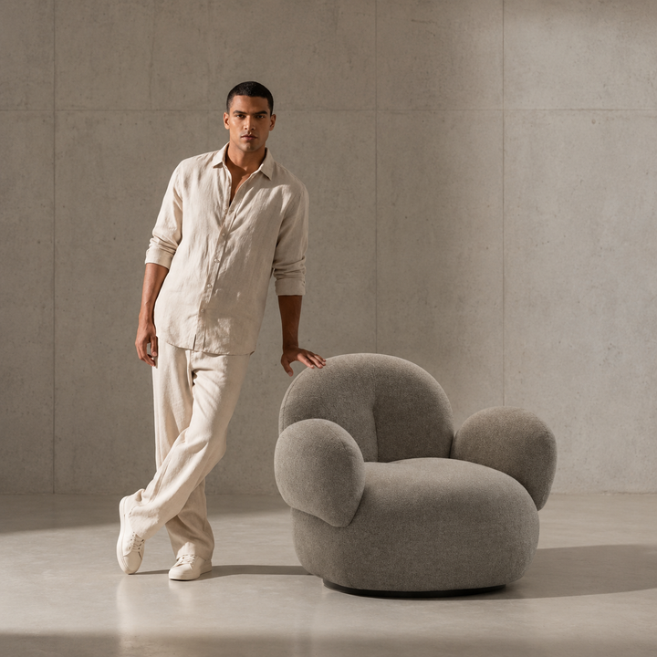 Man in a beige outfit standing next to a modern gray armchair against a neutral wall.