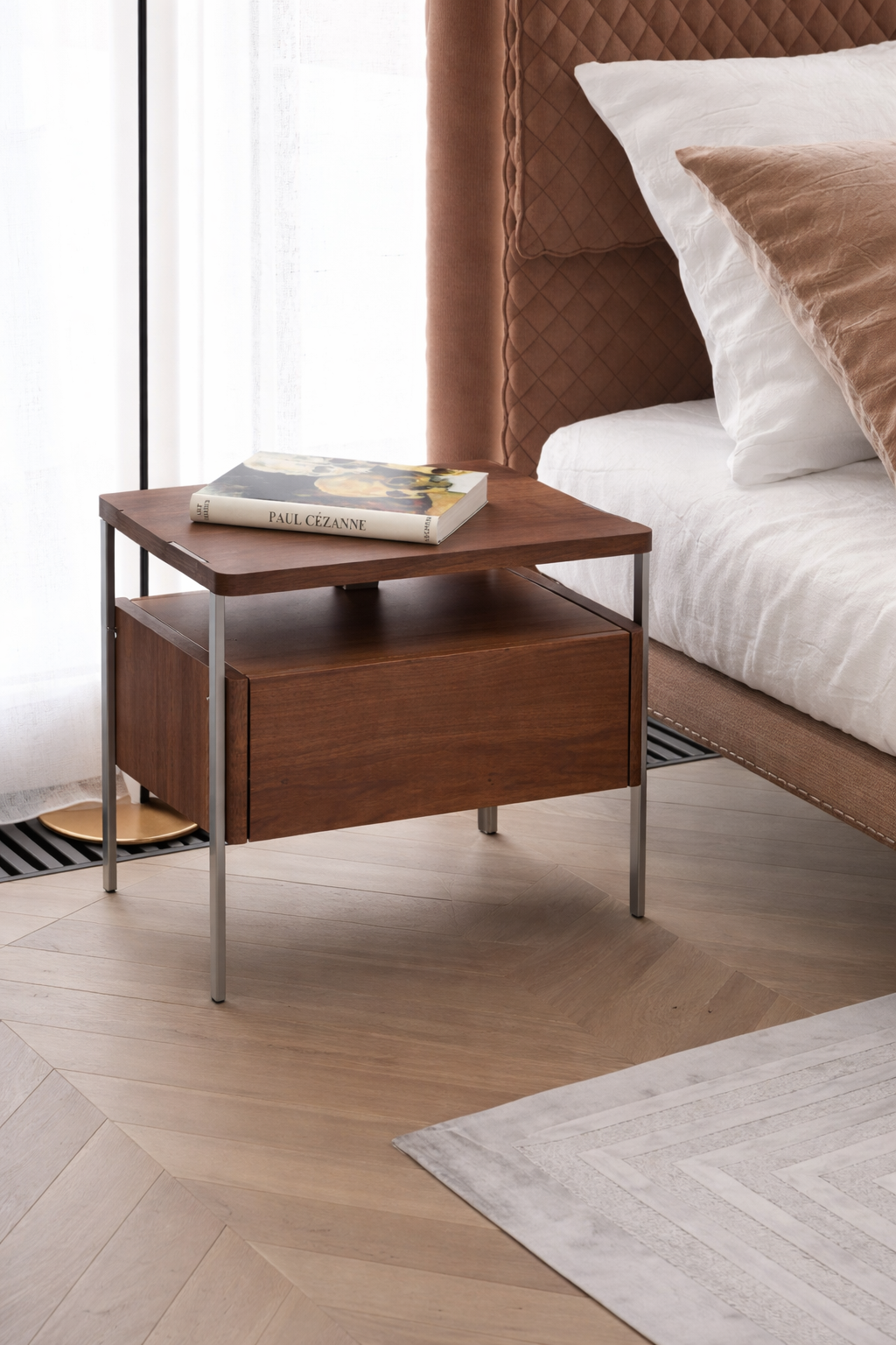 Wooden side table with a book next to a bed with white bedding and brown headboard.